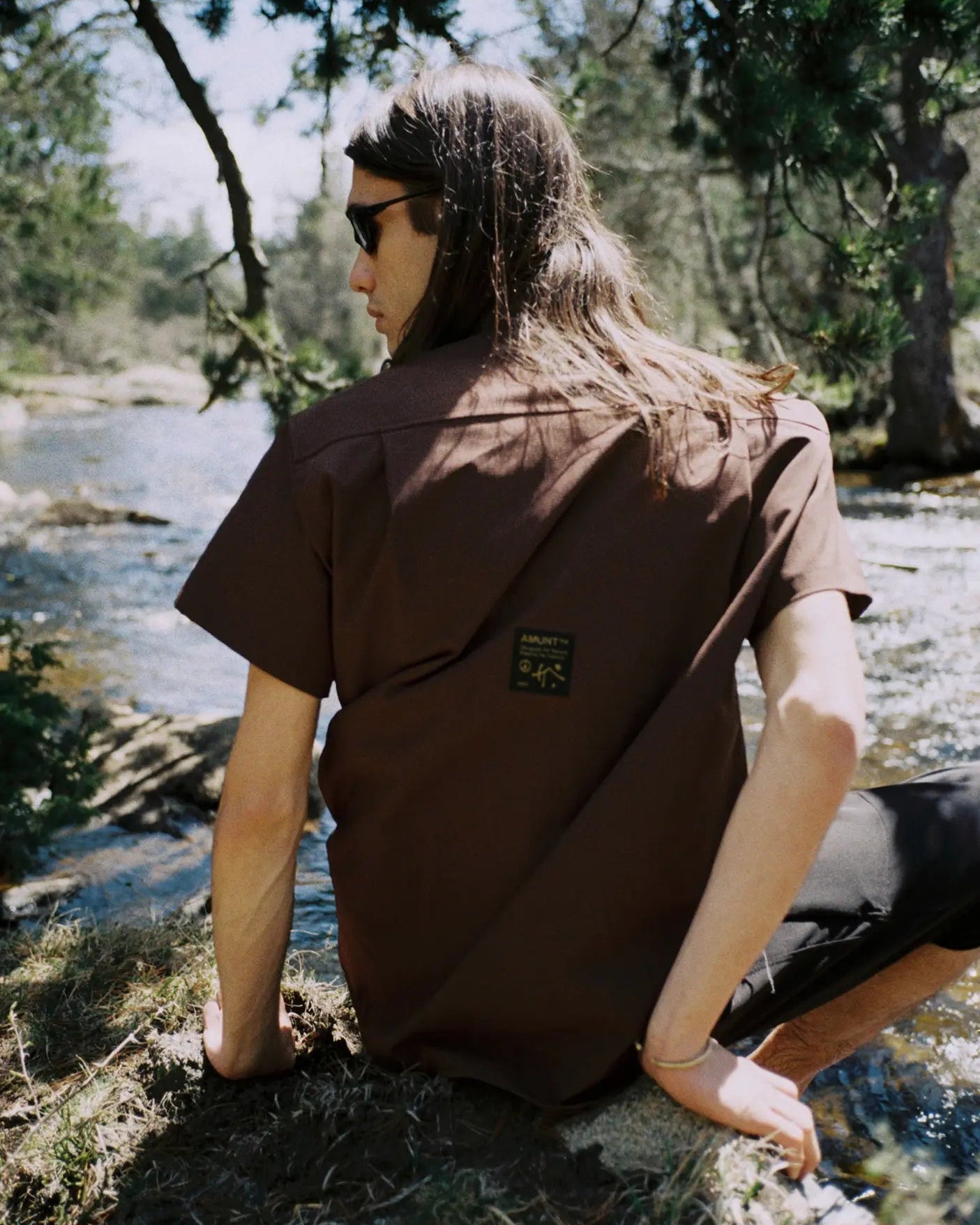 A person with long hair and sunglasses sits outdoors by a stream, wearing a brown short-sleeve button-up shirt with a small square patch on the back and dark pants.