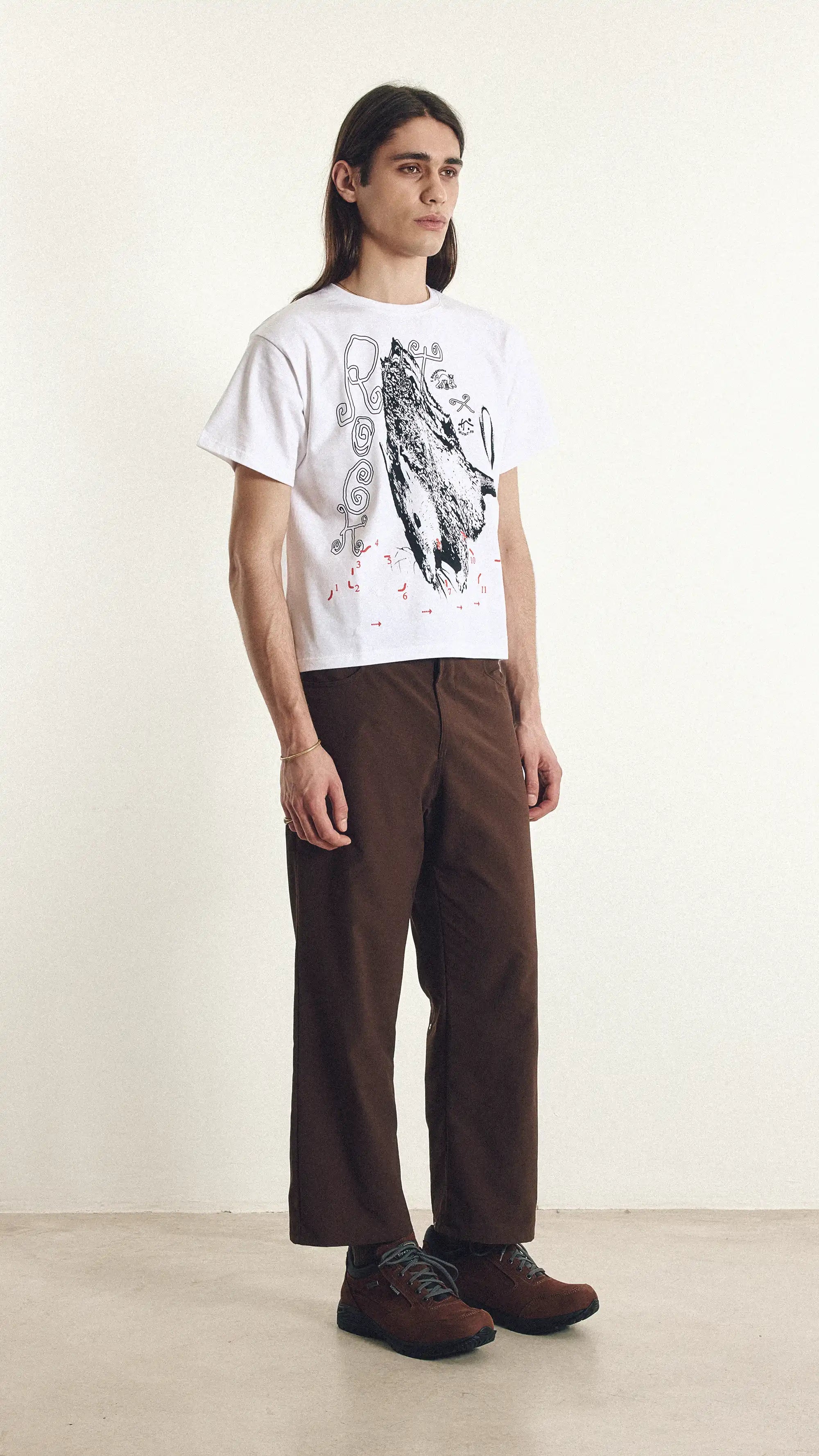 Person wearing a white t-shirt with a graphic design and brown pants on a plain background