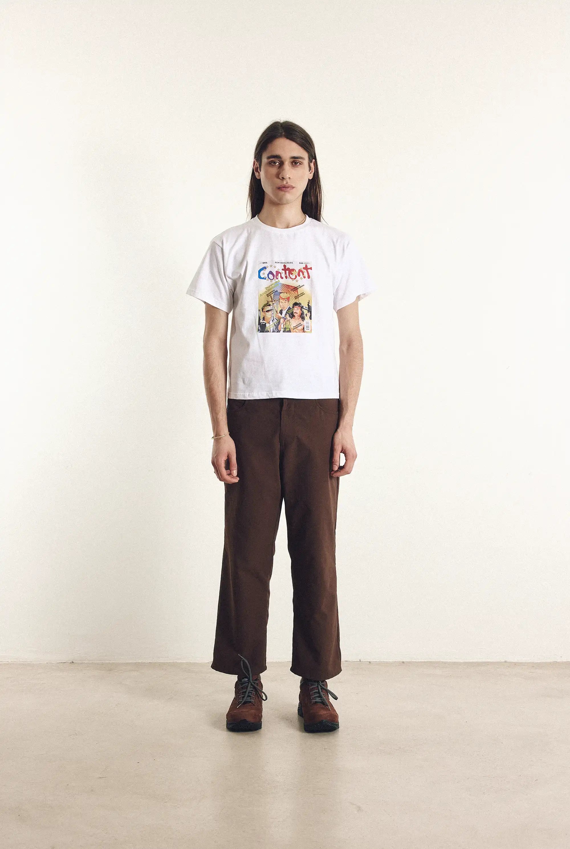 Person wearing a white t-shirt with colorful graphics and brown pants on a plain background
