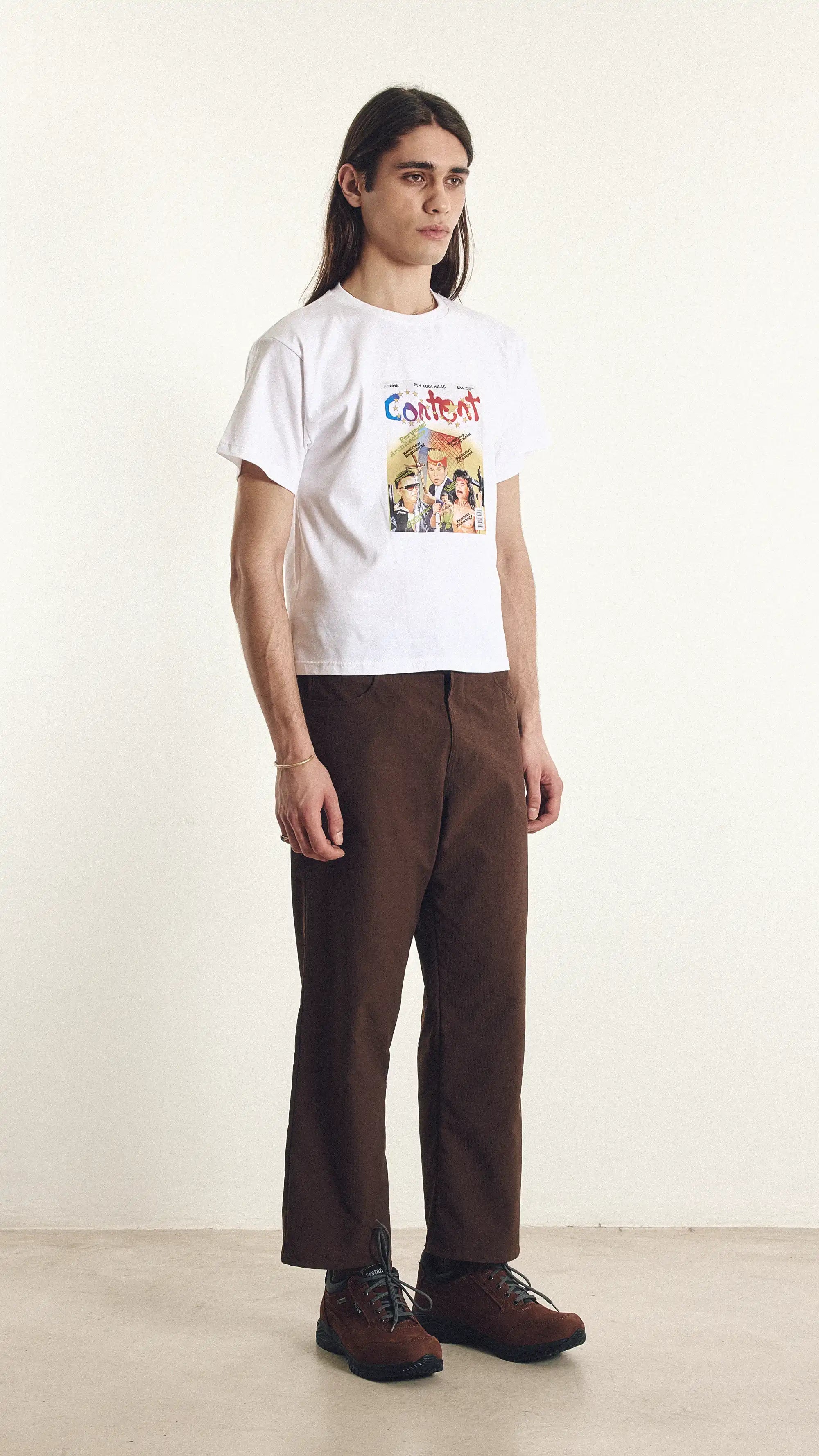 Person wearing a white t-shirt with colorful graphics and brown pants on a plain background