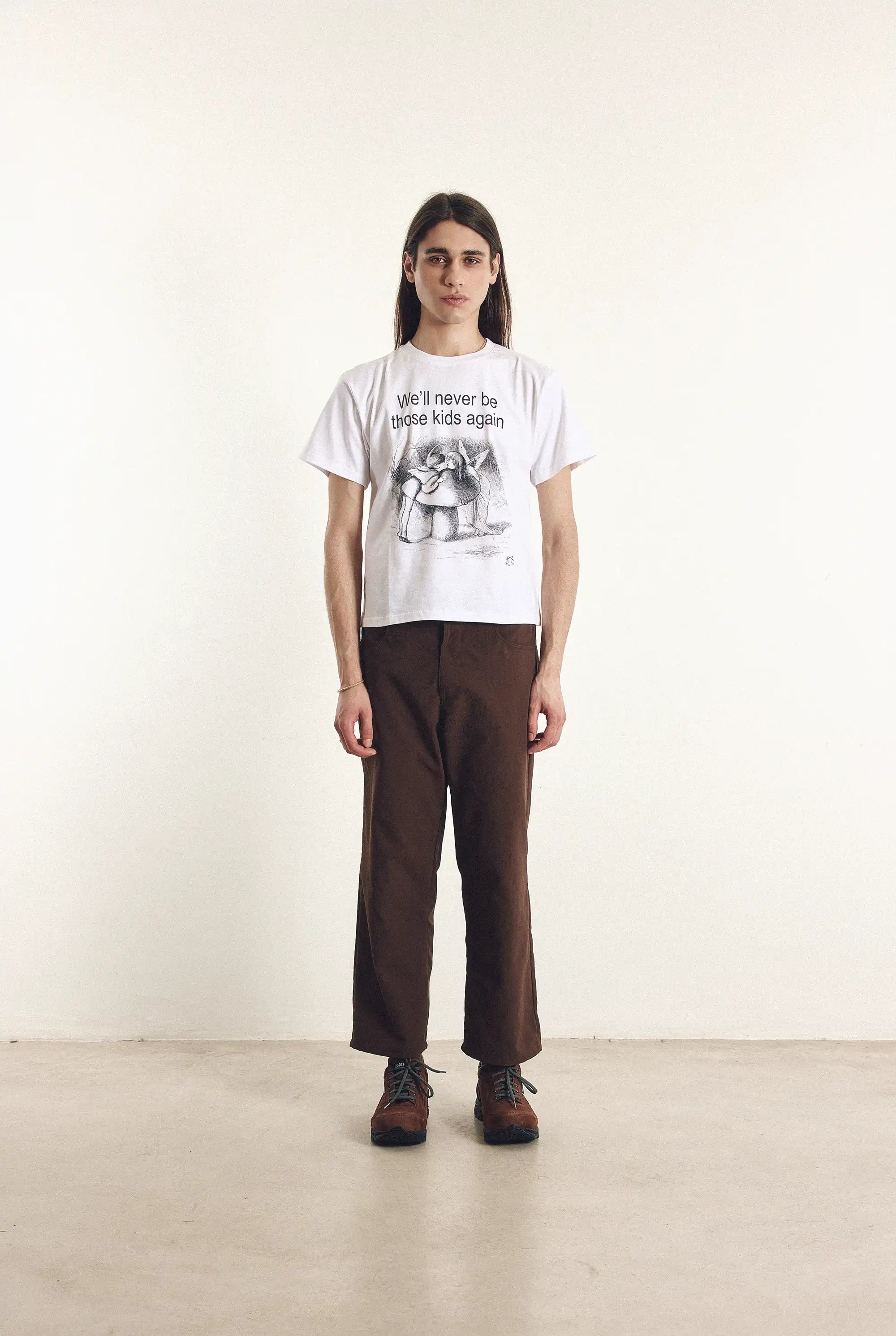 Person wearing a white t-shirt with text and graphics, brown pants, and dark shoes on a plain background