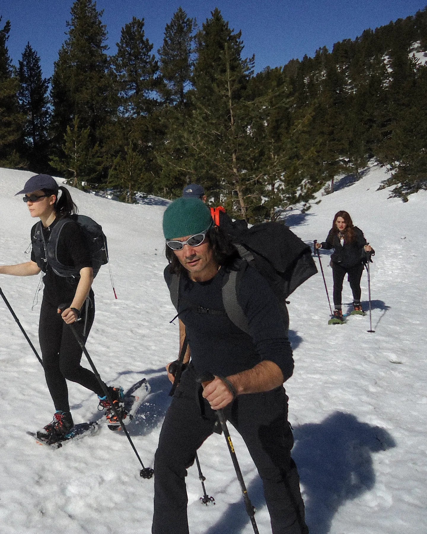Three people cross-country skiing on a snowy slope with trees in the background.
