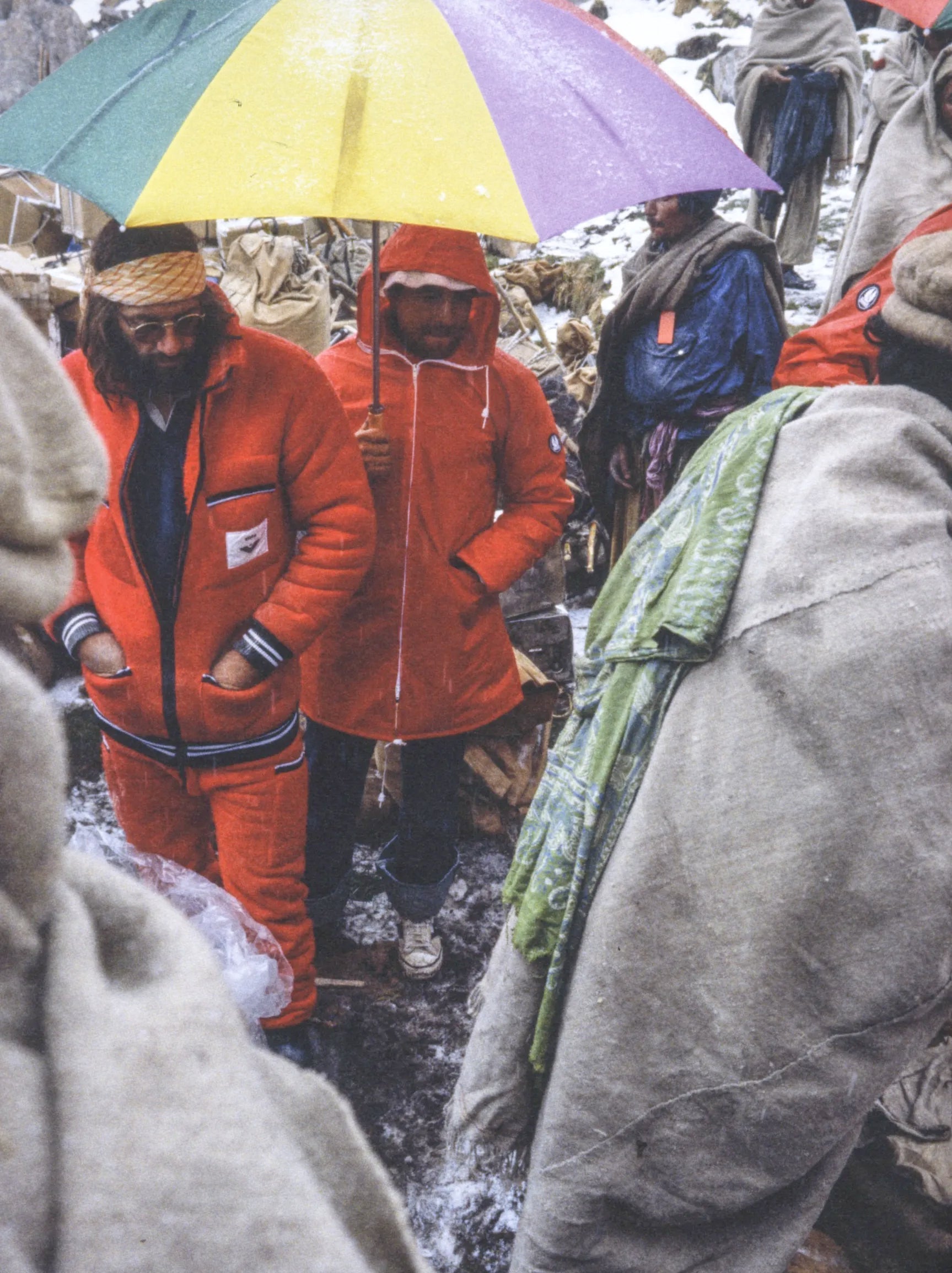 Person in orange snowsuit holding a colorful umbrella in a snowy landscape with other people.