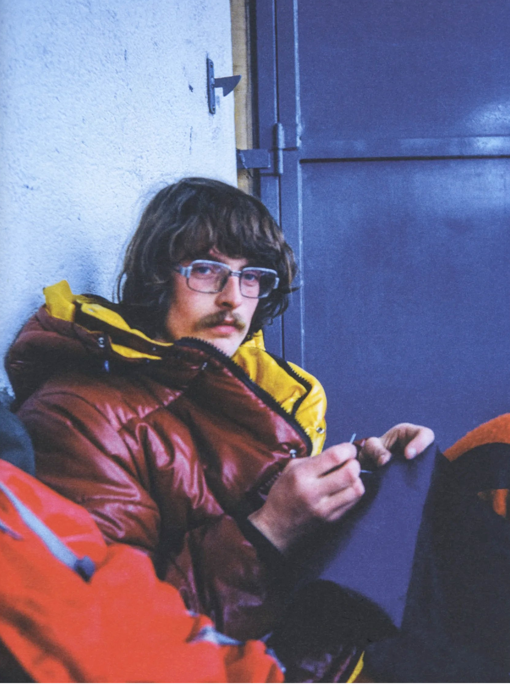 Person wearing a red jacket with a yellow hood, sitting against a blue wall.