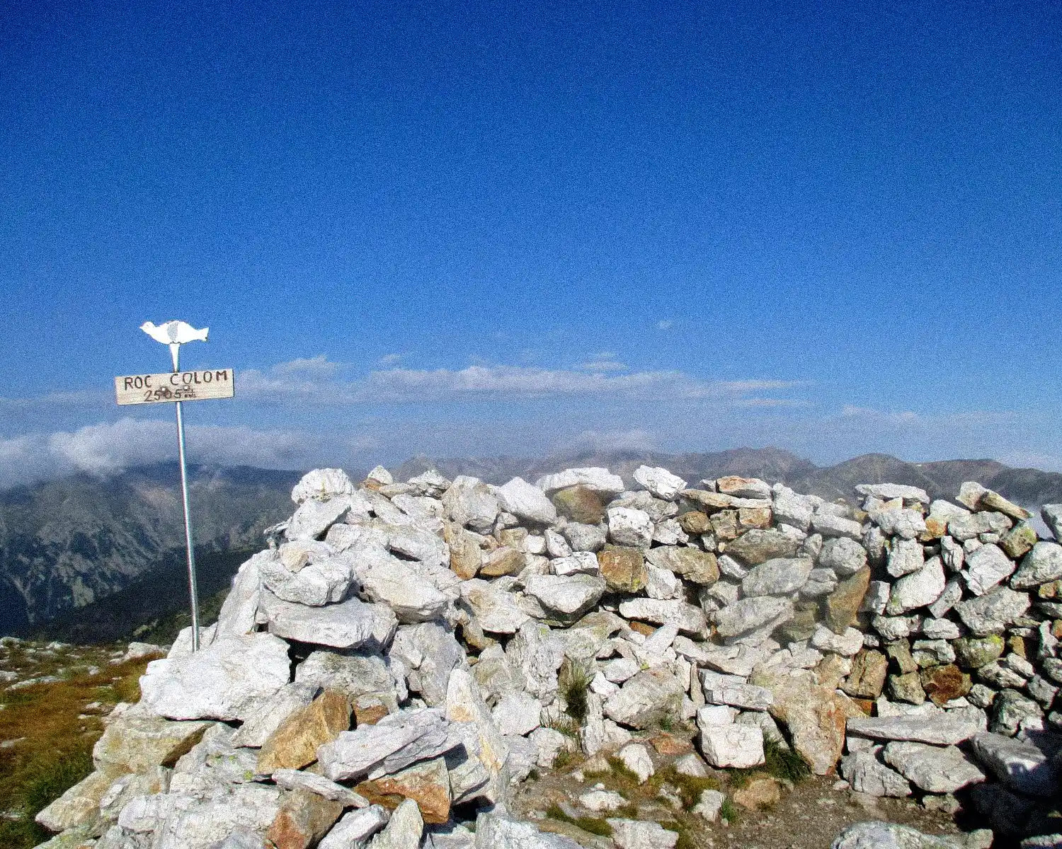 Stone cairn with a sign on top of a mountain peak under a clear blue sky.