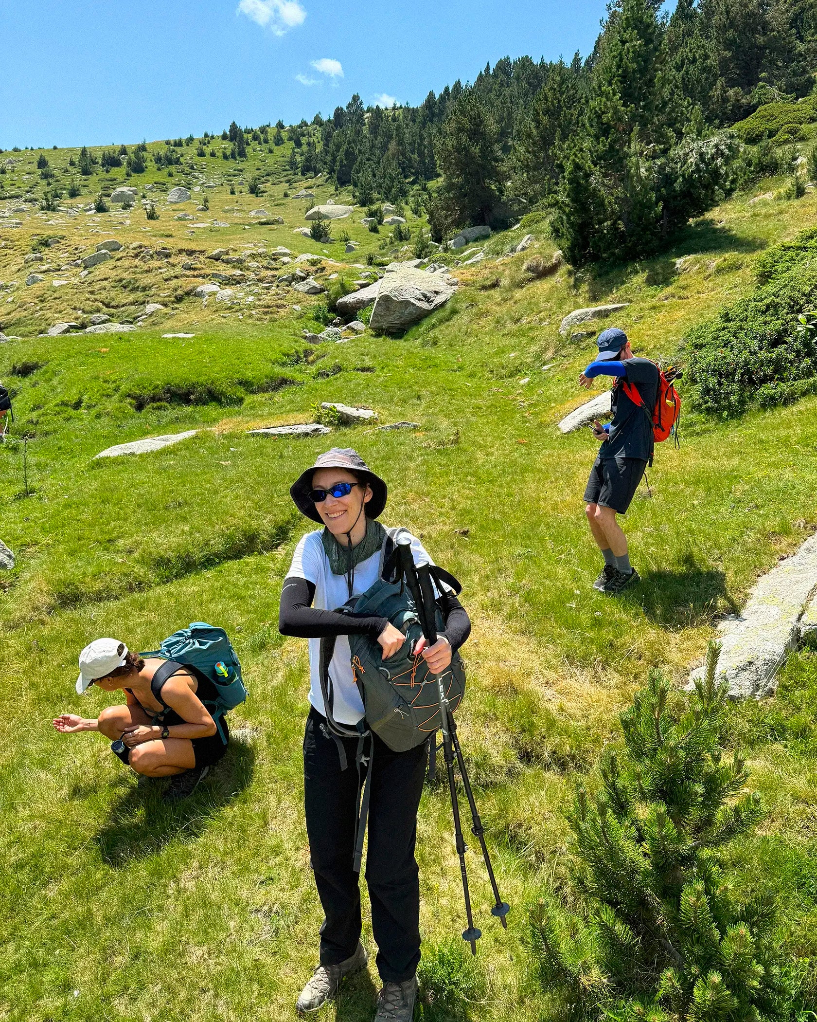 Three hikers on a grassy mountain slope under a blue sky. The central person wears a wide-brim hat, sunglasses, a light top, dark pants, and carries trekking poles and a backpack.