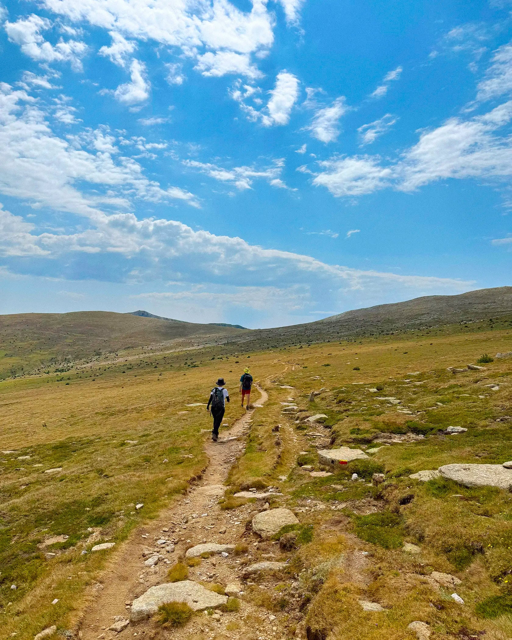Two hikers on a dirt trail in a grassy mountain landscape under a bright blue sky with scattered white clouds.