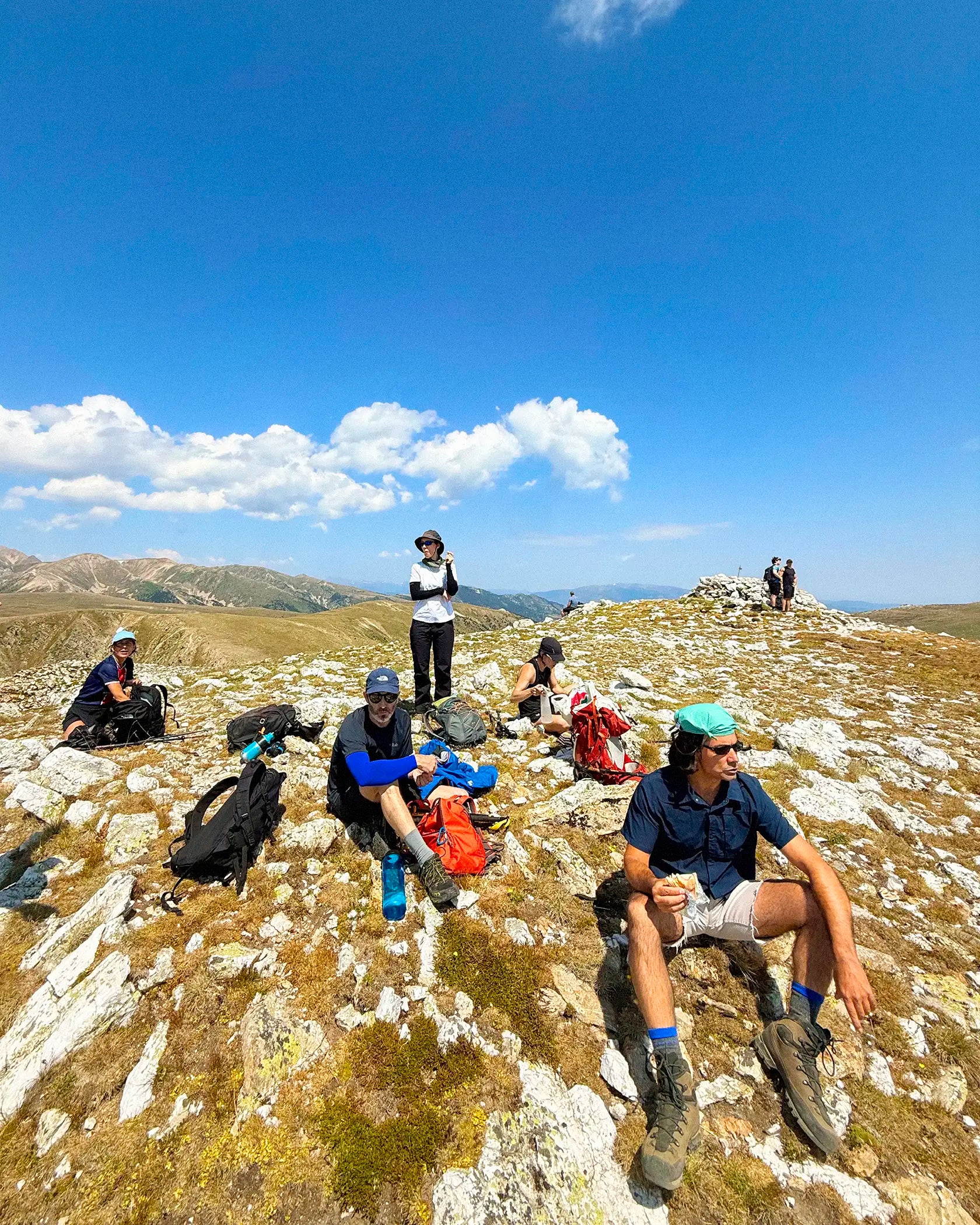 A group of hikers rests on a rocky, grassy mountain ridge under a bright blue sky with white clouds. In the foreground, a man in a navy shirt and green cap sits on a large white rock, while others are seated or standing further back with hiking backpacks and gear.
