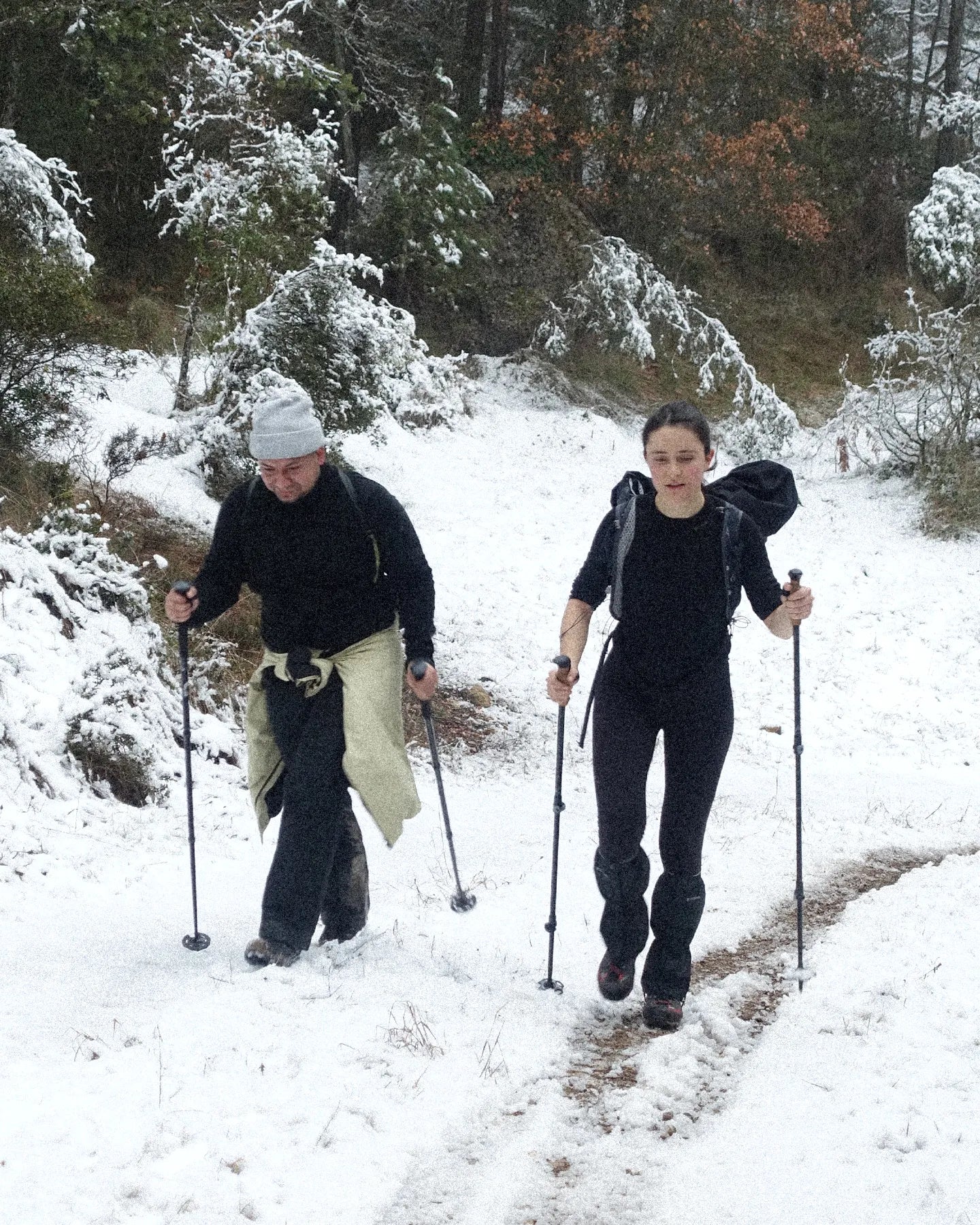 Two people hiking on a snow-covered trail with backpacks and walking sticks.