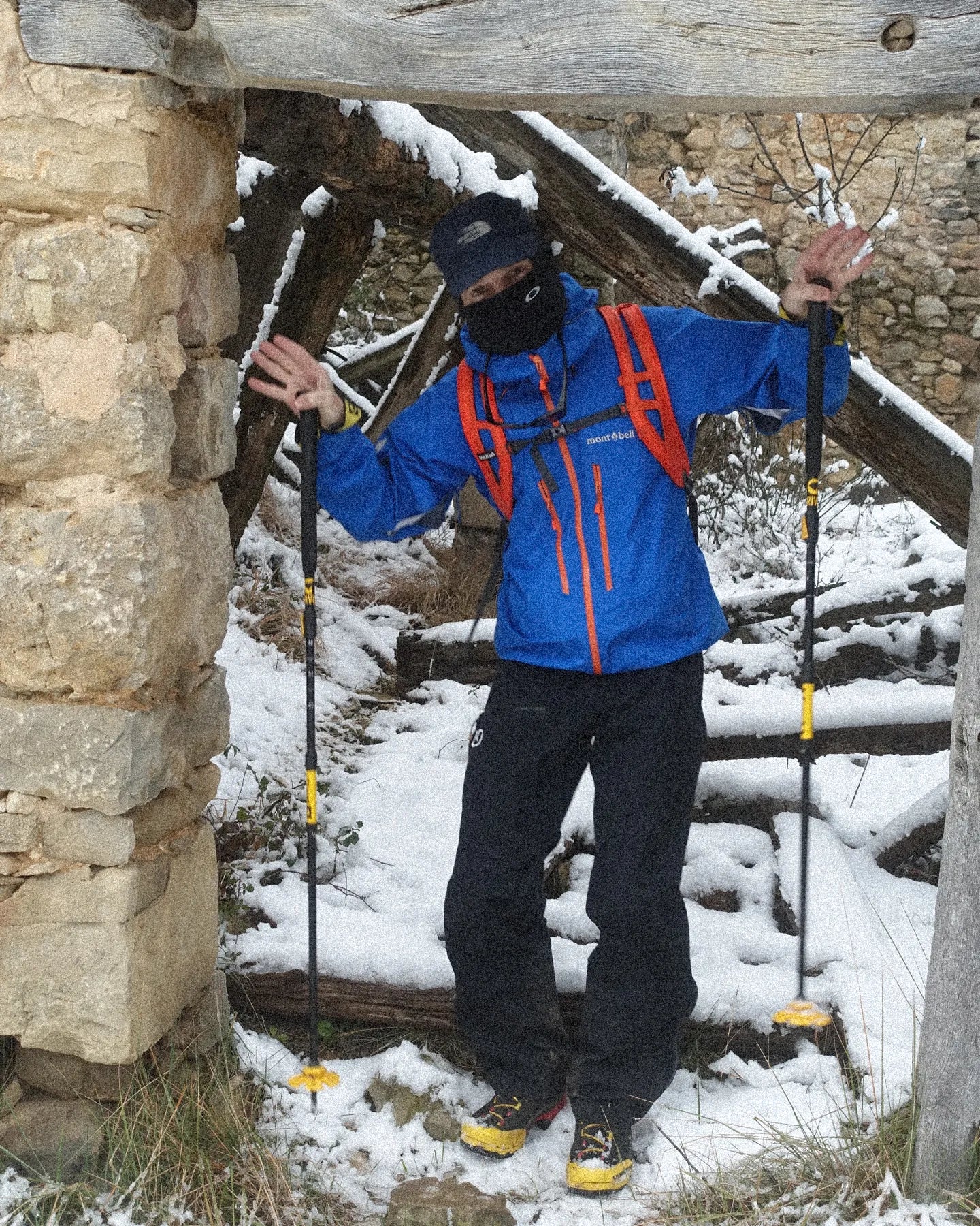 Person in blue jacket with orange accents standing on a snow-covered path with hiking poles.