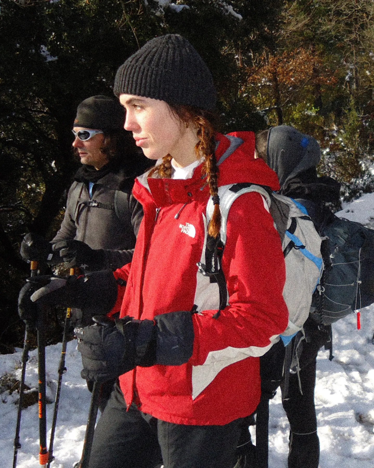 Two people hiking in a snowy landscape, one wearing a red jacket and black beanie, the other in dark clothing.